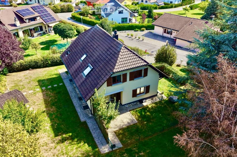 Aerial view of a single-family house with a brown tiled roof, light yellow walls, and multiple windows with brown shutters. The house is surrounded by a well-maintained garden with green lawns, trees, and a paved driveway leading to the entrance.
