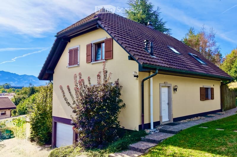 Two-story house with yellow walls, red-tiled roof, white windows with shutters, and a small garden with a tree and shrubs.
