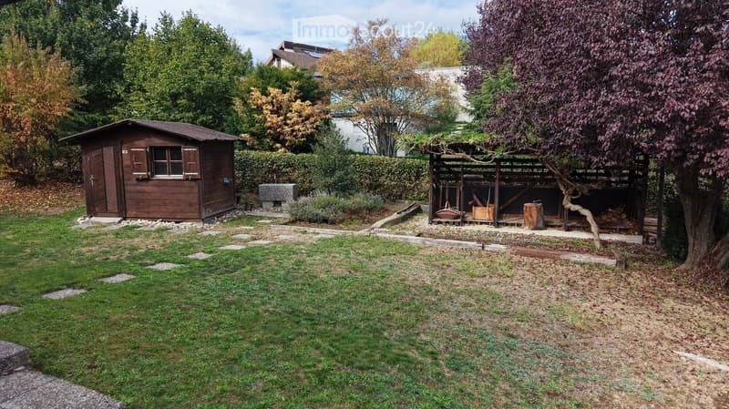 A well-maintained garden with a wooden shed, a tree, a stone bench, and a covered area with a wheelbarrow and logs.