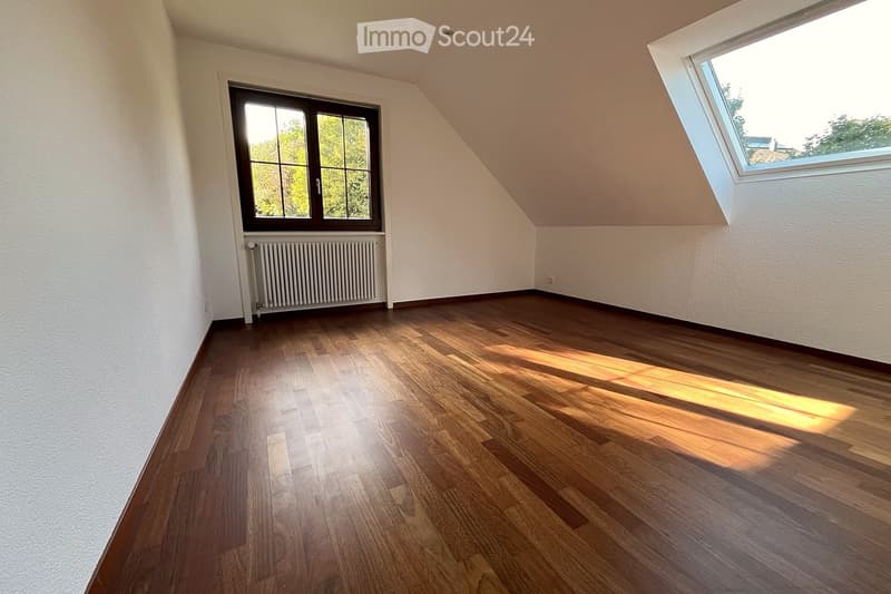 Empty room with wooden flooring, white walls, and large windows allowing natural light. Features a radiator and a skylight.