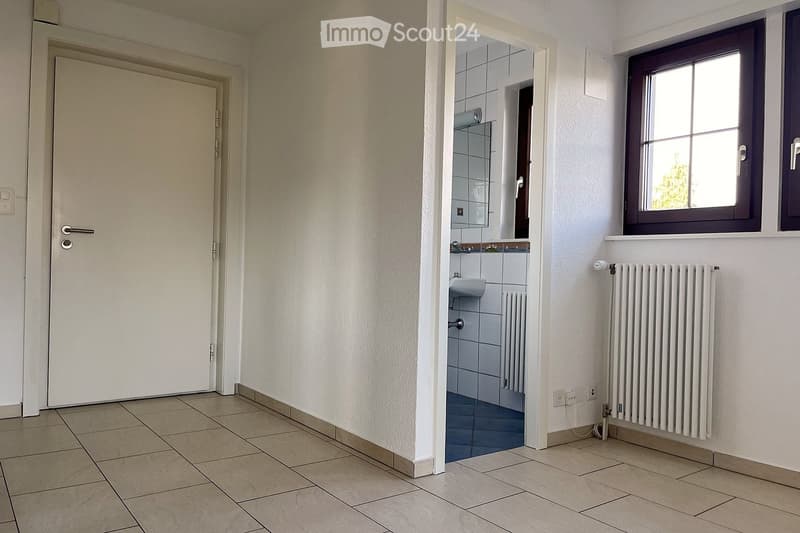 Empty hallway with tiled flooring, white walls, a door leading to a bathroom, and a window with a radiator below it.