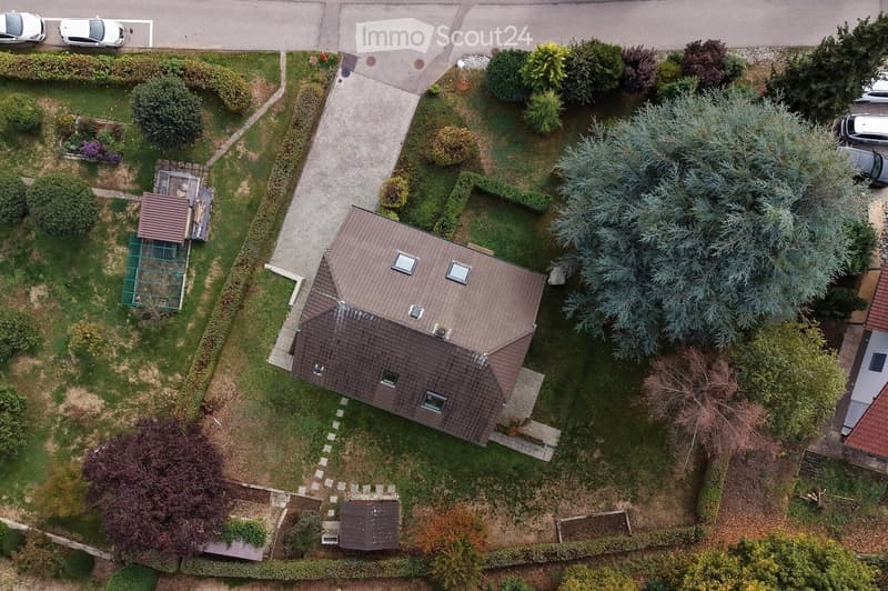 Aerial view of a two-story house with a brown tiled roof, surrounded by a well-maintained garden with various trees and shrubs. The house has multiple windows and a small garage. There is a paved driveway leading to the house, and several parked cars are