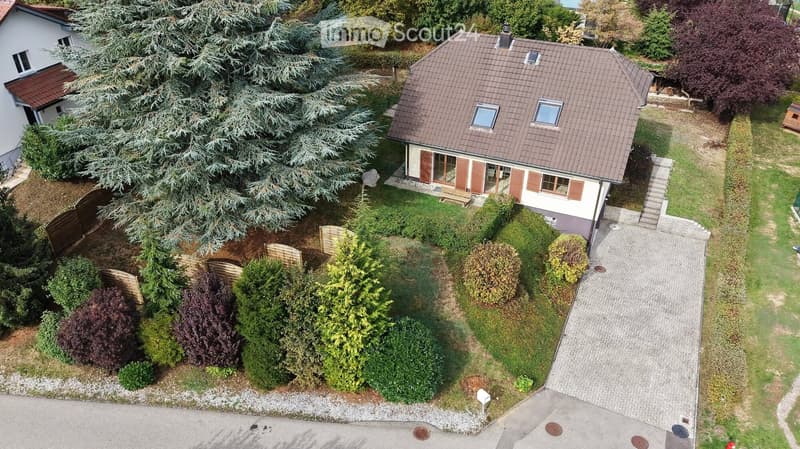 Aerial view of a two-story house with a brown tiled roof, surrounded by a well-maintained garden with various trees and shrubs. The house features multiple windows and a small balcony area.