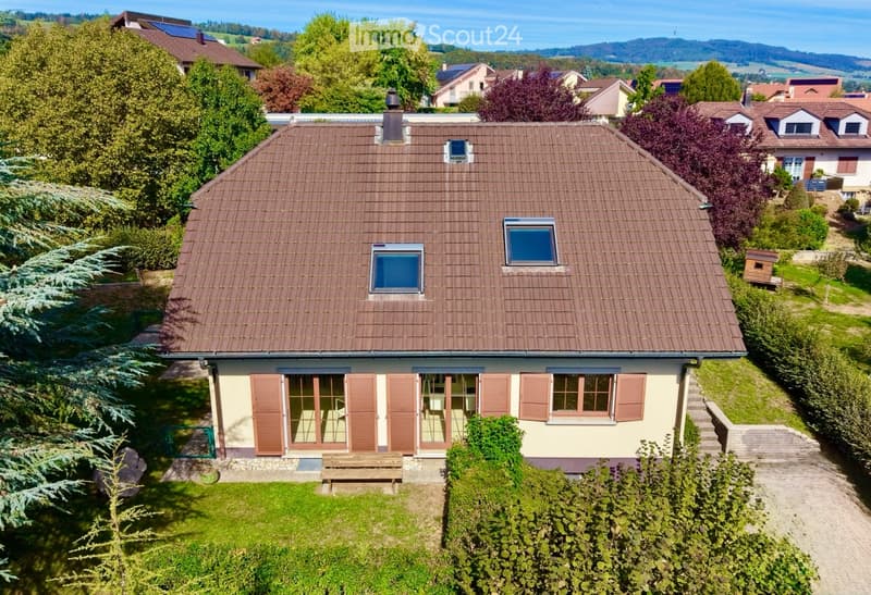 A two-story house with a brown tiled roof, multiple windows, and a well-maintained garden. The house features a light-colored facade with wooden shutters and a small porch area with a bench.