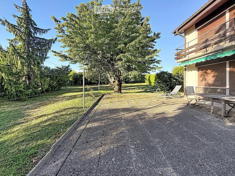 Garden area with gravel pathway, lounge chairs, and a table near a house with a balcony and awning.