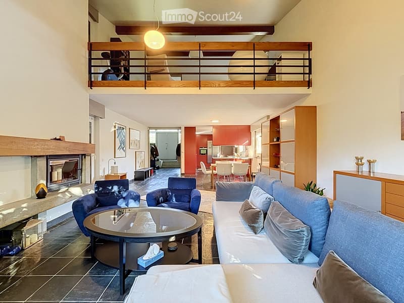 Modern living room with a fireplace, a large sofa, coffee table, armchairs, and a dining area with chairs and a table. Shelves and a small bar area are also visible.