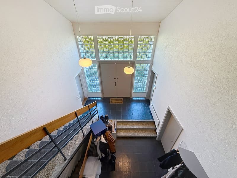 Spacious hallway with a staircase leading upwards, wooden railing, pendant lights, and a double door at the end.
