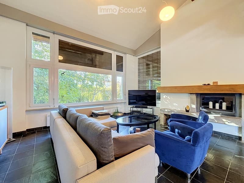 Modern living room with large windows, a grey sectional sofa, a blue armchair, a glass coffee table, a flat-screen TV, and a fireplace with decorative candles.