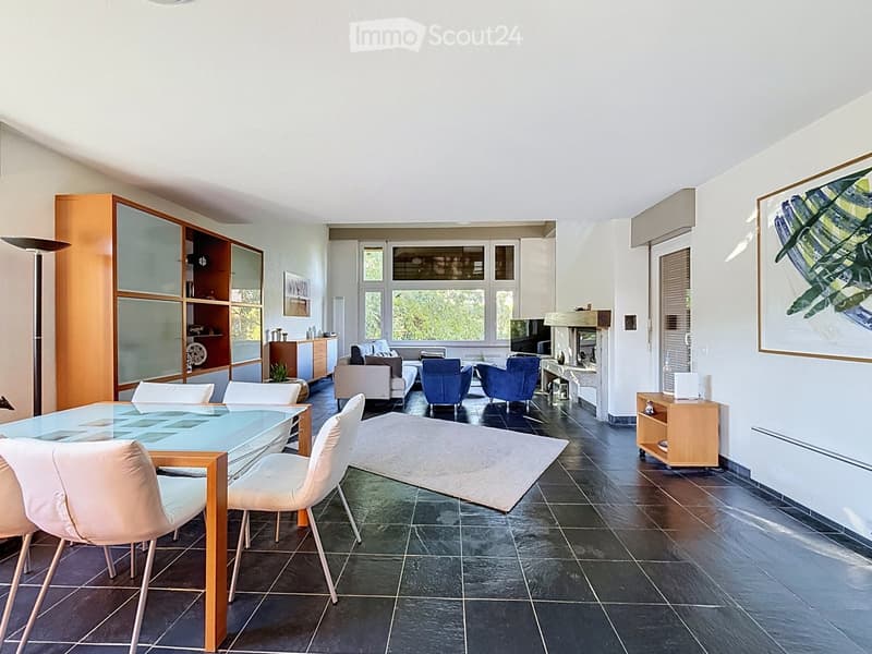 Modern living room with large windows, dark tiled floor, white sofa, blue armchairs, glass dining table, and wooden shelves.