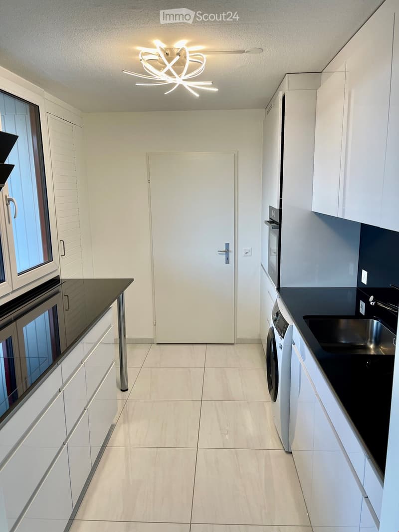 Modern kitchen with white cabinets, black countertops, a stainless steel sink, an oven, and a washing machine. The space features a large window and a contemporary light fixture.