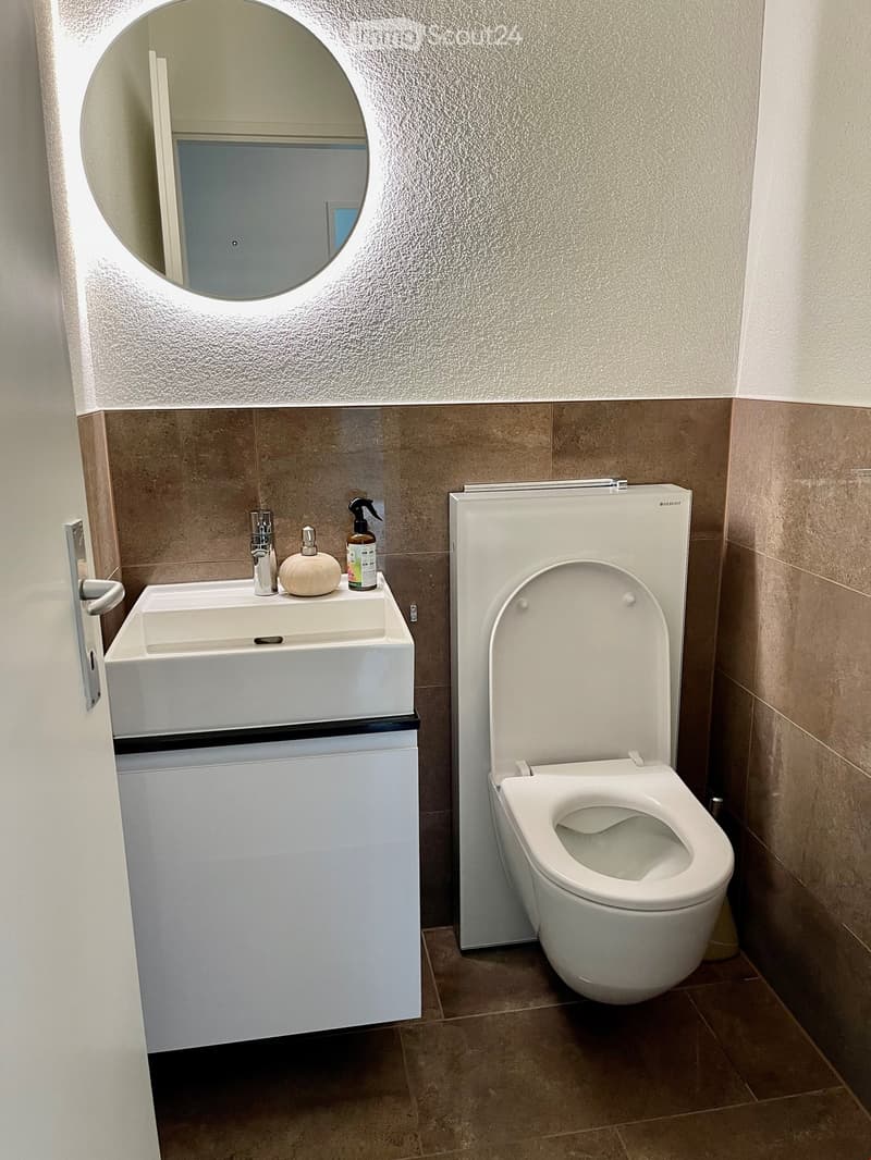 Modern bathroom with a white sink, chrome faucet, toilet, and a round mirror above the sink. The walls are tiled in beige and white, and the floor is tiled in brown.