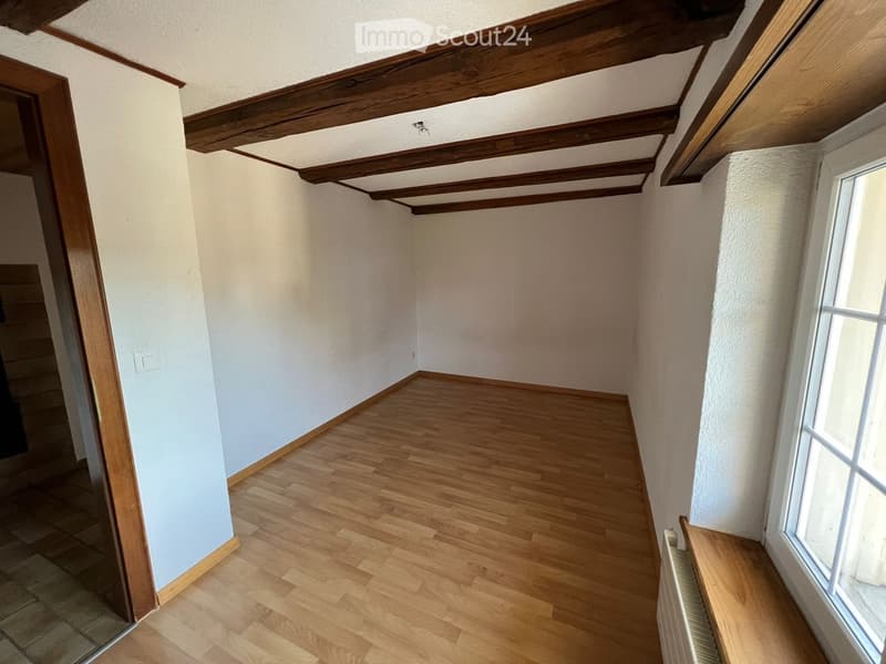 Empty room with wooden flooring, white walls, and exposed wooden beams on the ceiling. A window with white frames is visible on the right side.