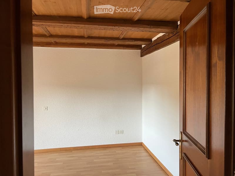 Empty room with light wooden flooring, white walls, and exposed wooden ceiling beams. A wooden door is visible on the right side.