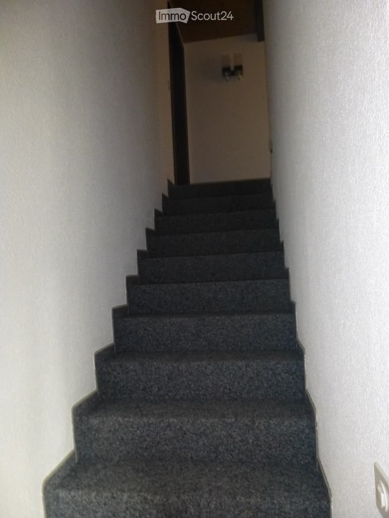 A narrow staircase with dark, carpeted steps and white walls, leading upwards.