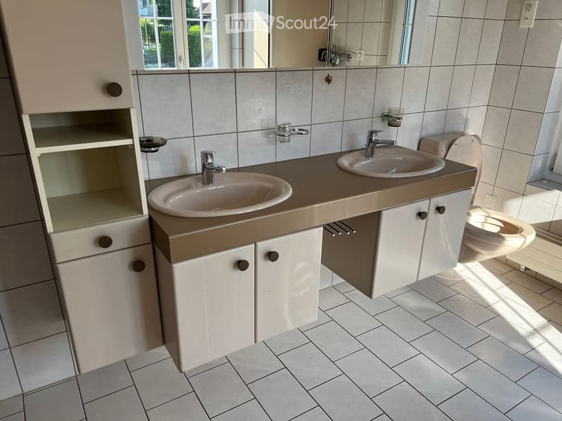 Modern bathroom with two sinks, a toilet, and white cabinets with black handles. The floor is tiled, and the walls are tiled with a light gray color. There is a large mirror above the sinks.