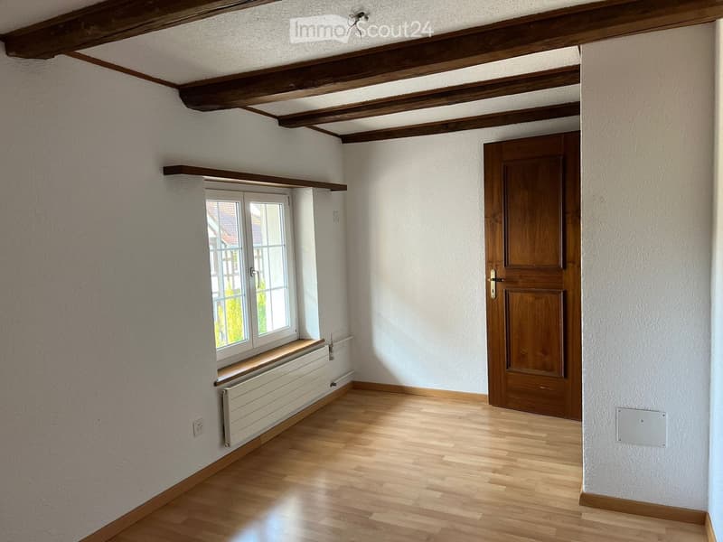 Empty room with wooden flooring, white walls, exposed wooden beams on the ceiling, a window with a built-in seat, and a wooden door.