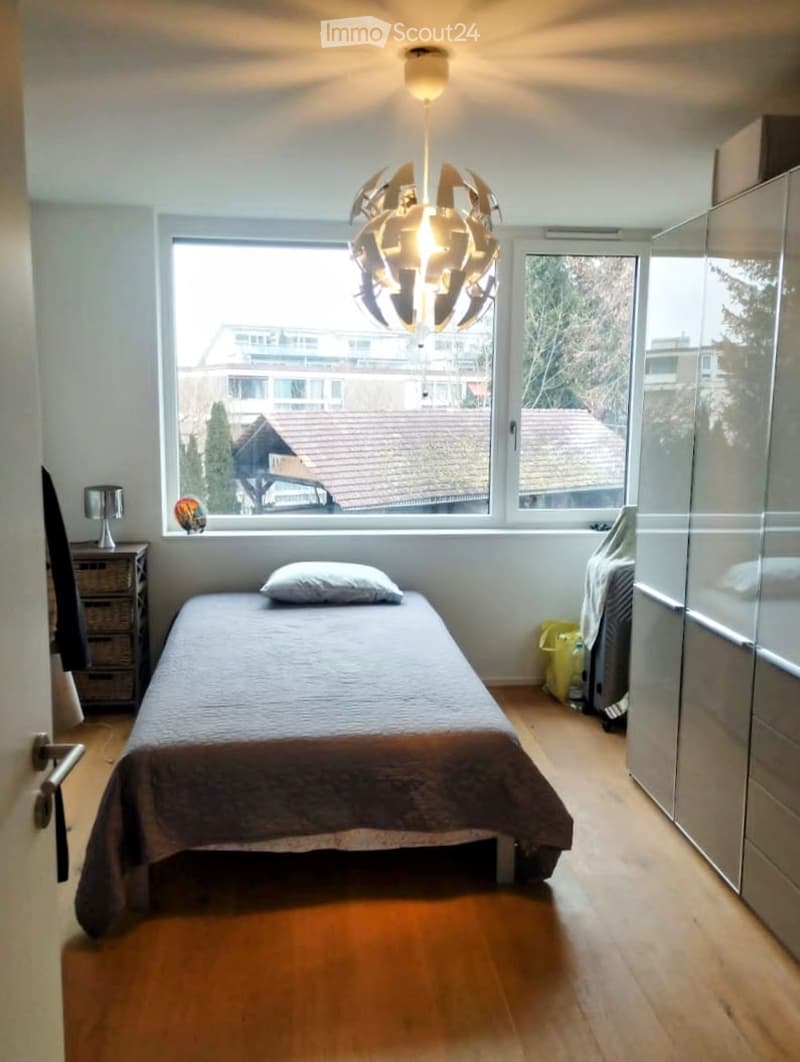 Bright bedroom with wooden flooring, large windows offering a view of neighboring houses, a single bed with a pillow, a modern hanging light fixture, a wardrobe, and a nightstand with a lamp.