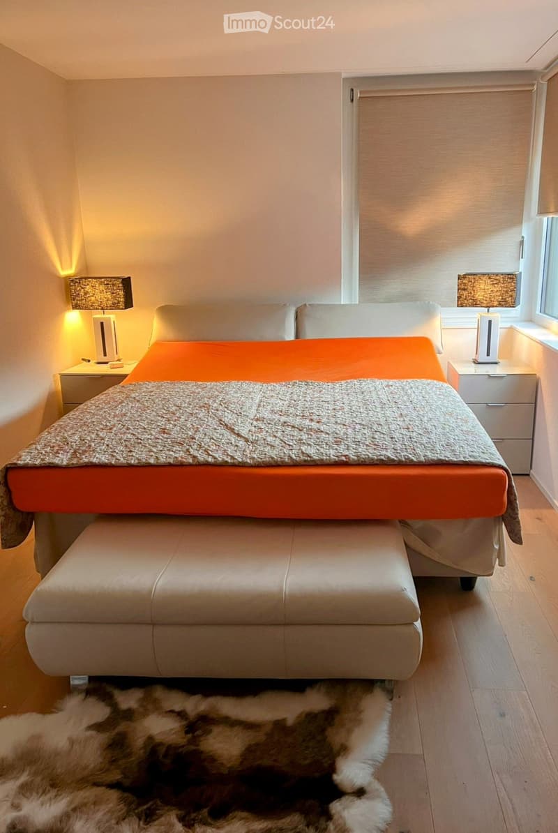 Modern bedroom with a large bed, orange and grey bedding, a leather stool at the foot of the bed, nightstands with lamps, and a fur rug on the floor.