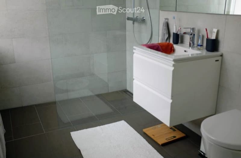 Modern bathroom with a white sink cabinet, a mirror, a shower with a glass door, a toilet, a towel, and a bathroom scale on the floor.
