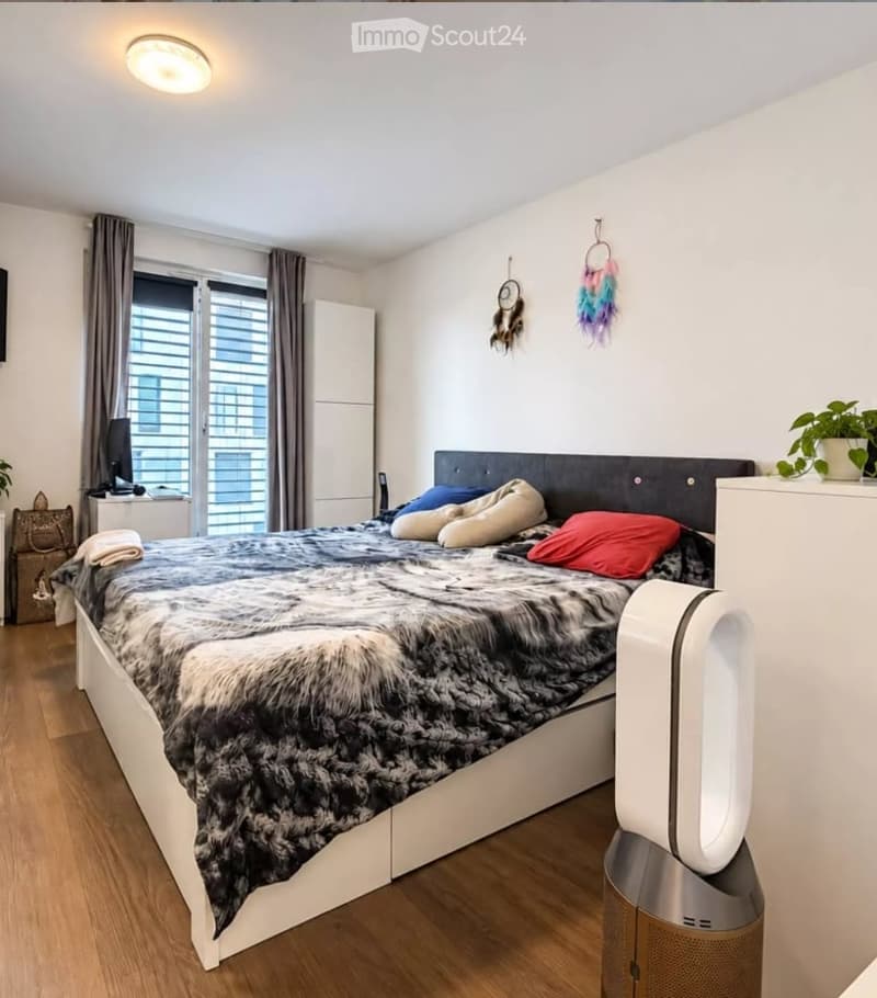 A modern bedroom with a large bed covered in a fur-like bedspread and colorful pillows, a nightstand with a screen, a fan, and decorative items on the walls.