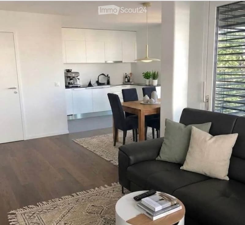 Modern open-plan living and dining area with a dark leather sofa, coffee table, dining table with chairs, and a sleek kitchen area with white cabinets and a coffee machine.