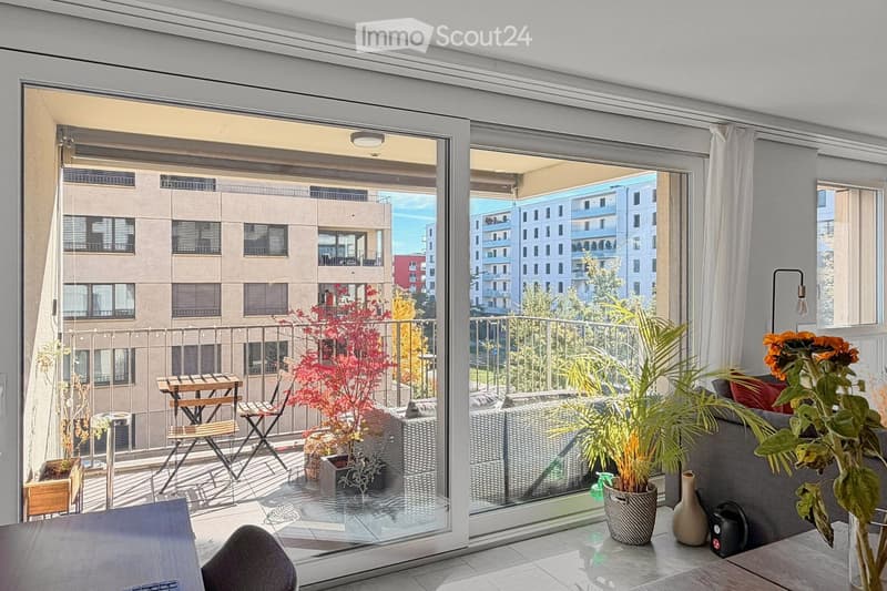 Modern living room with large glass doors opening to a balcony, featuring a grey sofa, wooden chairs, a small table, and decorative vases with plants.