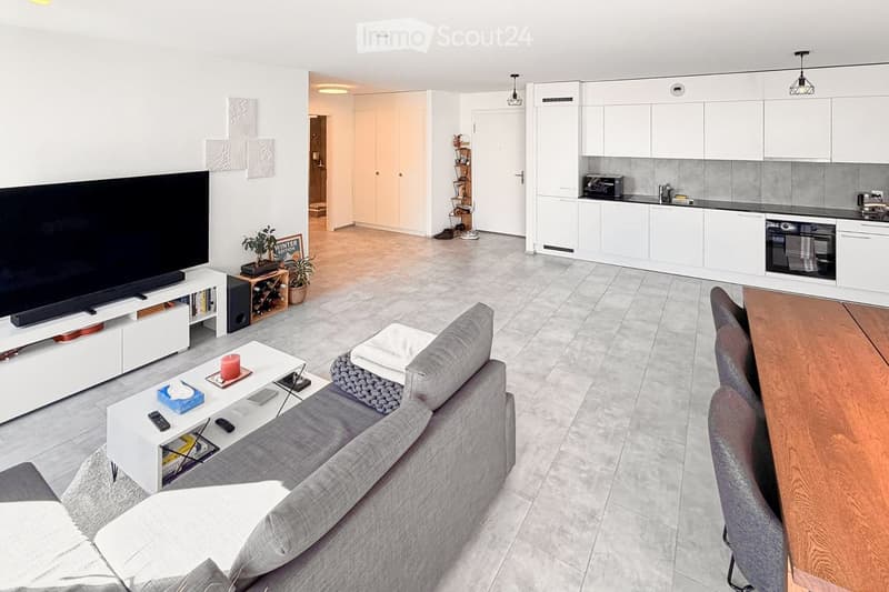 Modern living room with grey sofa, white coffee table, TV stand, and a small kitchen area with white cabinets and an oven. The space includes a wooden dining table with chairs, a wine rack, and decorative plants.