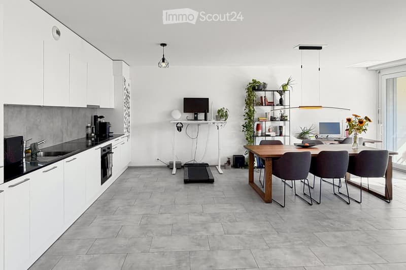 Modern kitchen and dining area with white cabinets, grey tiles, and a wooden dining table with four chairs. A laptop and screen are on a desk, and there are plants and a vase with flowers on the table.
