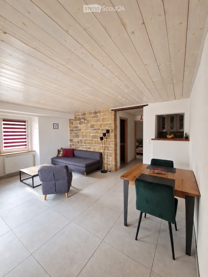 Bright living and dining area with a slanted wooden ceiling, white tiled floor, grey sofa, coffee table, armchair, dining table with chairs, and a floor lamp near a stone pillar.