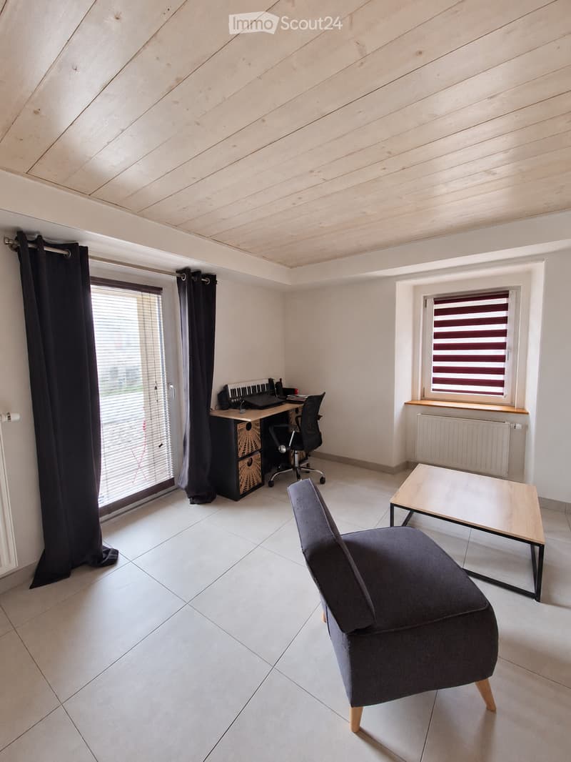 Bright room with wooden slatted ceiling, white tiled floor, large windows with blinds, a dark grey sofa, a small wooden table, a desk with a laptop, and a black office chair.