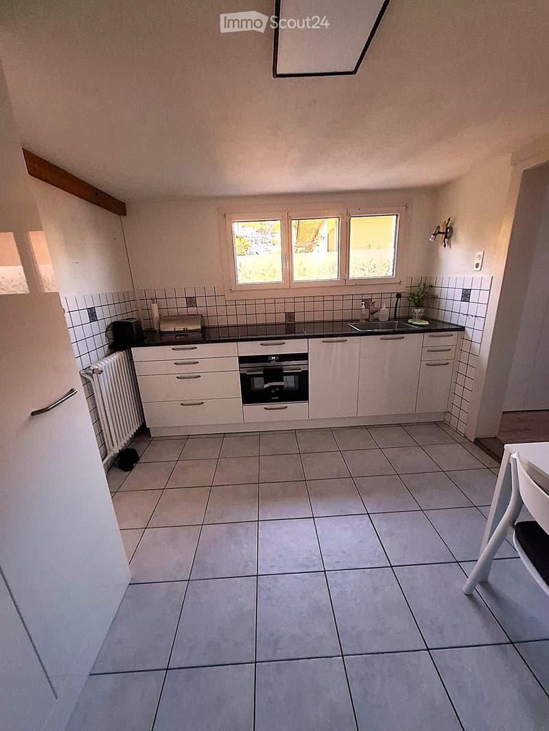 Modern kitchen with white cabinets, tiled backsplash, and stainless steel appliances. Features an oven, sink, and radiator, with a small chair visible in the foreground.