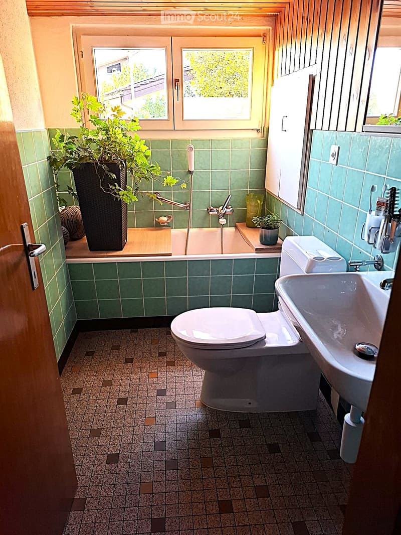 Bathroom with green tiled walls, a bathtub, toilet, sink, and a small plant on the windowsill.