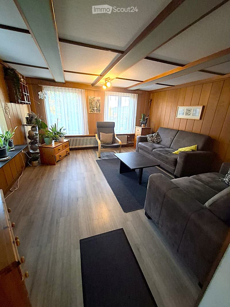 Cozy living room with wooden walls, grey sofa, coffee table, armchair, and potted plants on a side table. Large windows with white curtains let in natural light.