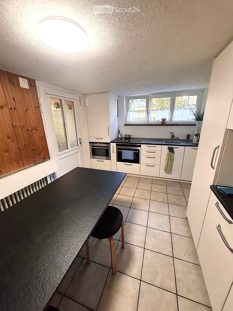 Modern kitchen with white cabinets, black countertops, and tiled flooring. Features a central island with a stool, built-in ovens, and a sink area with a window above.