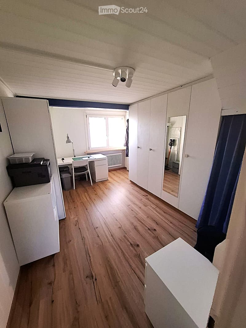 A furnished room with wooden flooring, a desk with a chair, a lamp, a mirror, a wardrobe, and a curtain on the window.