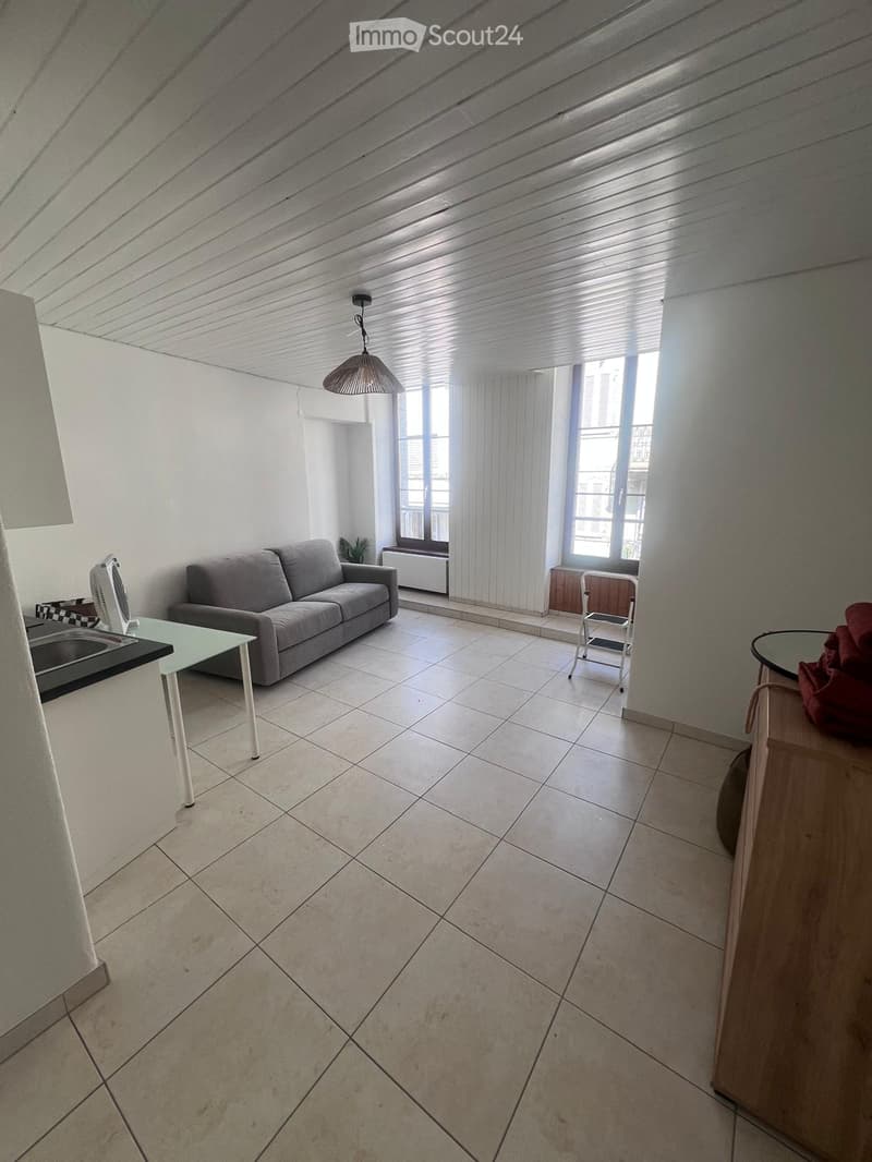 Bright living room with tiled floor, white walls, large windows with curtains, a grey sofa, a glass table, a fan, a small chair, and a mirror on a wooden stand.