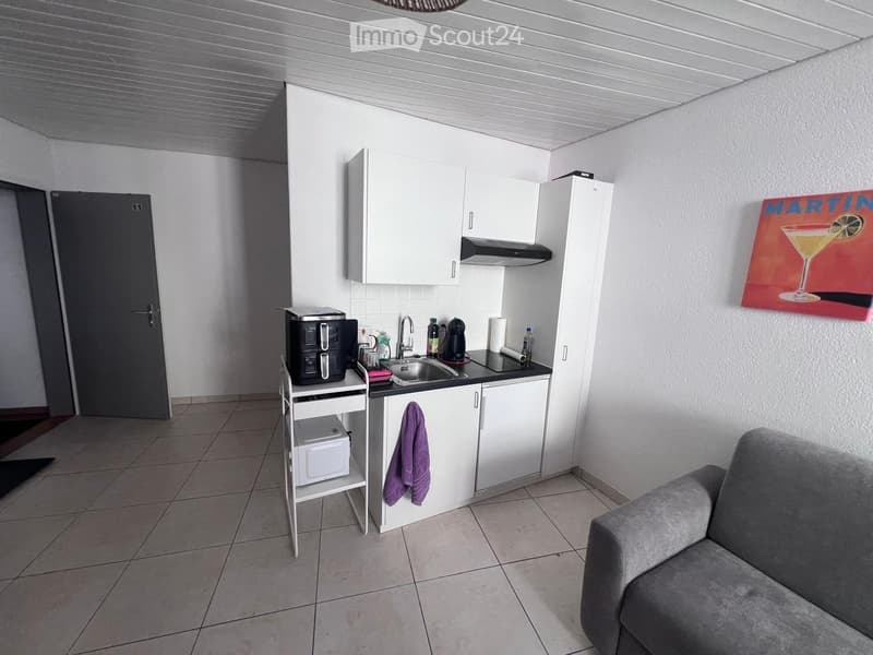 A compact, modern kitchen area with white cabinets, a sink, coffee maker, microwave, and a small sofa in the foreground.