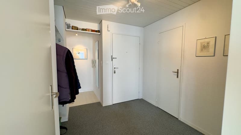 Clean, well-lit hallway with carpeted floor, white walls, and two closed doors. A coat rack with jackets is visible on the left, along with a small shelf holding decorative items and framed artwork.