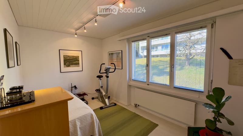 Well-lit room with large windows, exercise bike, small table with items, framed pictures on the wall, and a potted plant near the window.