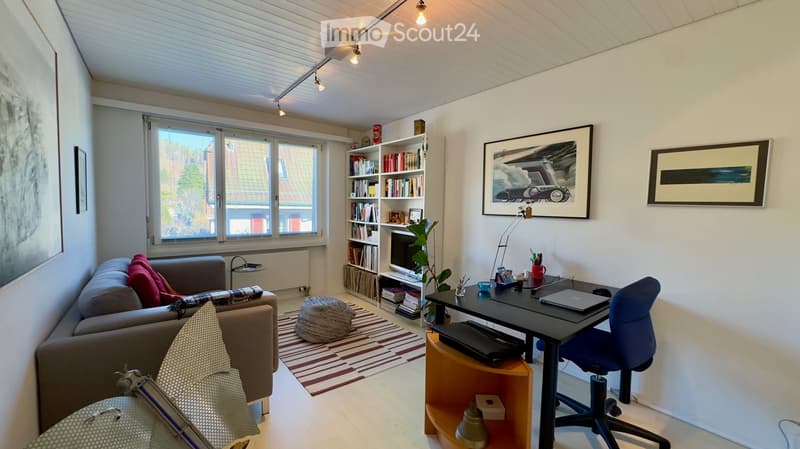 A modern, well-lit living room with a sofa, a desk with a laptop, a bookshelf, and a small plant. The room features large windows with a view of the outside, a patterned rug, and several decorative items including picture frames and a table lamp.