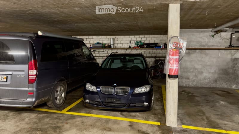 Underground parking area with two parked vehicles, a van and a car, concrete floor, and metal shelving with various items.