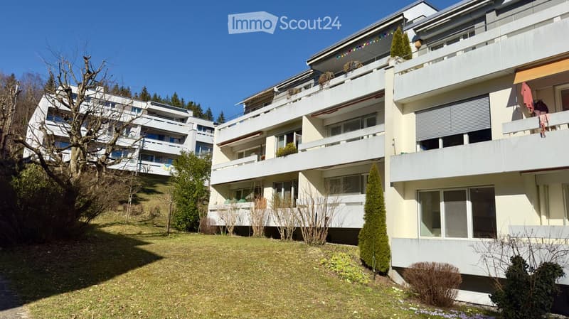 Multi-story residential building with balconies, surrounded by greenery and a grassy area.