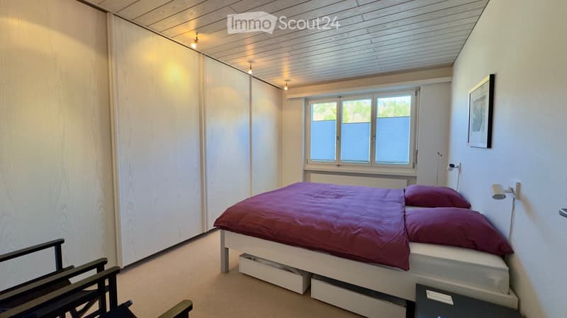 Modern bedroom with a double bed, dark red duvet, two pillows, a wooden chair, a nightstand, and a framed picture on the wall. The room features light wood paneling on the walls and ceiling, a large window with blinds, and built-in storage units.