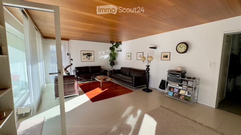 Modern living room with light wooden flooring, white walls, and large windows. Features a dark leather sofa, a coffee table with a red rug, an armchair, a floor lamp, a record player on a metal shelf, and a wall clock.
