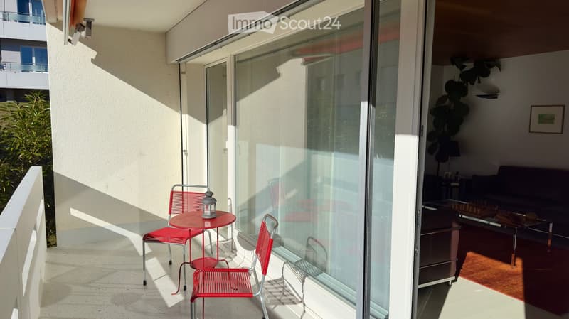 Balcony with a red metal table and chairs, lantern, and a view into a living room with a sofa and coffee table.