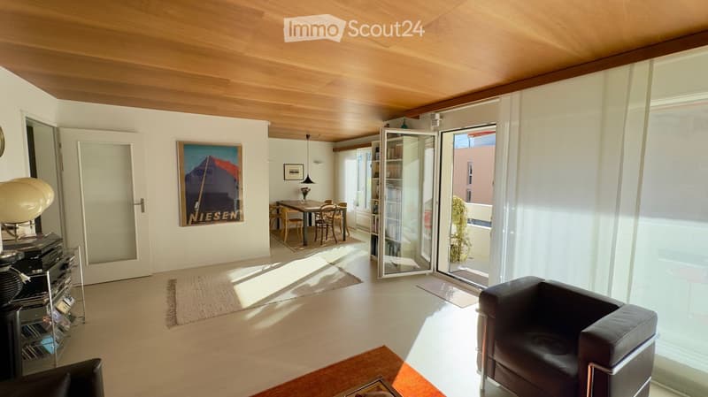 Bright living room with wooden floors, large windows, and a balcony door. Features a black leather sofa, a small dining area with a table and chairs, a bookshelf, and a decorative lamp.