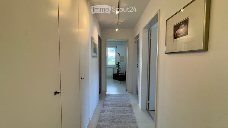 A clean, well-lit hallway with white walls, a gray rug, and framed artwork. The hallway leads to an open area with a small table and a potted plant visible in the distance.