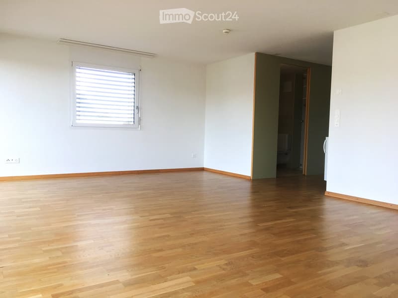 Empty room with wooden flooring, white walls, and a window with blinds. A doorway leads to another room.