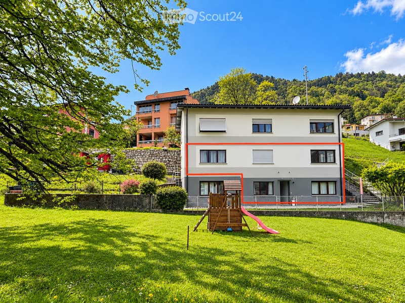 A two-story house with a modern design, featuring large windows and a balcony. The house is surrounded by a well-maintained lawn and trees, with a wooden play structure in the front yard.