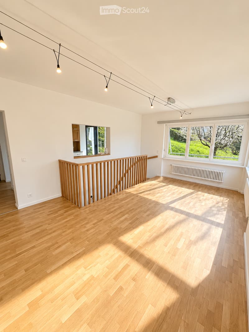Empty room with light wooden flooring, white walls, and large windows letting in natural light. Features a wooden railing and a small mirror on the wall.